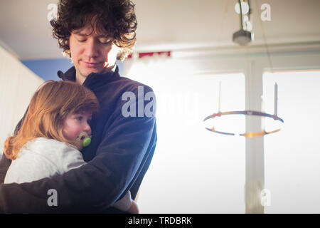 Young dad and toddler baby girl with pacificator embracing at home in natural morning light. Stock Photo