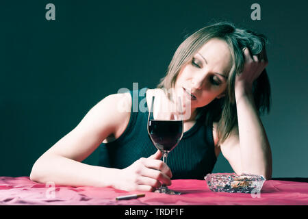 Portrait of a depressed and alcoholised woman holding a glass of wine with cigarettes in front of her Stock Photo