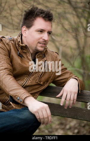 Portrait of a young likeable man dressed in a leather jacket sitting on a park bench Stock Photo