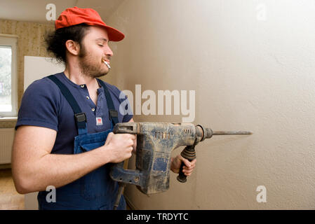 Workman dressed in a boiler suit holding a roto hammer to a wall Stock Photo