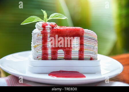 Crape cake slice with strawberry sauce on white plate and nature green background / Piece of cake rainbows with whipped cream Stock Photo