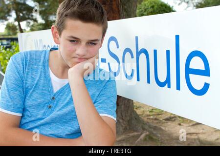 Angry looking boy sitting outside in front of a school sign Stock Photo