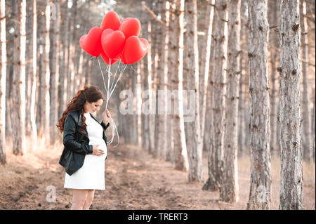 Pregnant woman 30-35 year old holding heart shape red balloons standing in forest. Wearing black leather jacket and white dress outdoors. Maternity. M Stock Photo