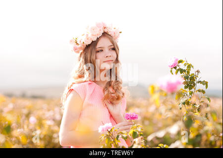 Smiling teenage girl 14-16 year old wearing pink dress and floral wreath with roses standing in rose field. Stock Photo