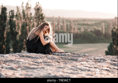 Blonde teen girl 14-16 year old sitting alone on rock over beautiful nature background. Looking at camera. Loneliness. Stock Photo