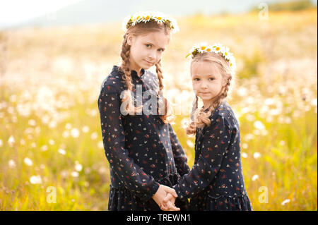 Two girls holding hands standing in chamomile meadow. Looking at camera. Wearing stylish vintage dresses. Childhood. Stock Photo