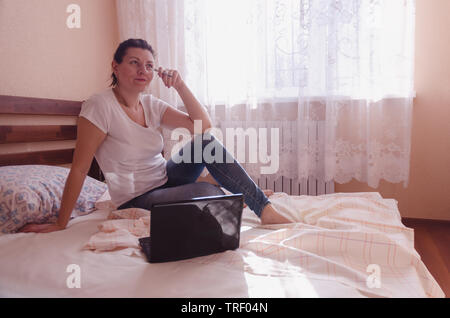 A happy young woman works on a laptop in a cozy bedroom in the sunny morning. Stock Photo