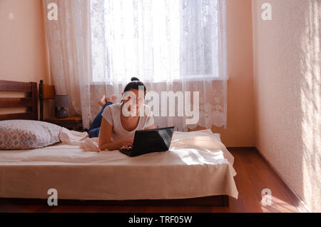 A happy young woman works on a laptop in a cozy bedroom in the sunny morning. Stock Photo