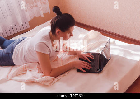 A happy young woman works on a laptop in a cozy bedroom in the sunny morning. Stock Photo