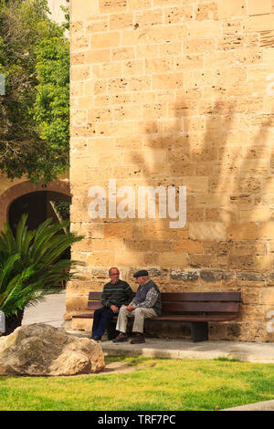 Two elderly gentlemen chatting on a bench next to the medieval walls of Alcudia old town in Majorca. Stock Photo