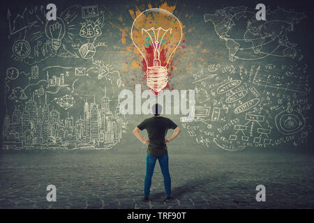 Rear view of confident young man, hands on hips, stands in front of a wall with drawn colorful light bulb and business sketches. Uniqueness of the ide Stock Photo