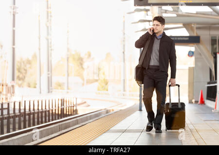 Businessman business traveler with roller bag suitcase talking on cell phone on train station platform Stock Photo