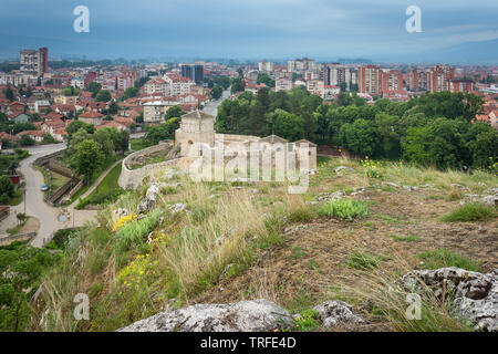 Low angle shot with foreground rocks and vivid yellow flowers in a foreground and ancient fortress and Pirot cityscape at the distance during blue hou Stock Photo
