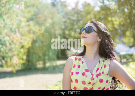 Beautiful long-haired brunette in a light silk dress with polka dots  and sunglasses rejoices in the sun against the backdrop of summer nature Stock Photo