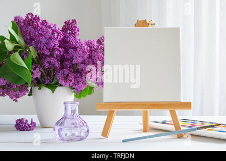 Blank canvas on easel, watercolor paints, brush for painting and lilac flowers on table. Stock Photo