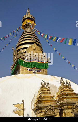 The gold spire and all-seeing eyes of Buddha atop a white-domed stupa festooned in prayer flags, Swayambhunath Buddhist temple, Kathmandu, Kathmandu V Stock Photo