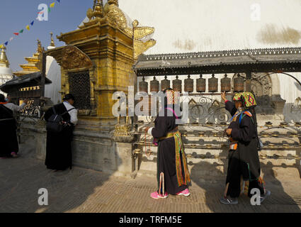 Tibetan pilgrims in traditional dress spinning prayer wheels and worshipping at a gilded niche of the large stupa at Swayambhunath Buddhist temple, Ka Stock Photo