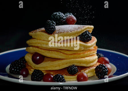 Stacked pancakes with summer berries and falling powdered sugar, isolated on black. Stock Photo