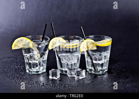 Three glasses of fresh cold carbonated water with ice cubes and lemon slices. Soda water on black background. Stock Photo