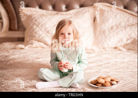 Cute child girl 3-4 year ol having breakfast in bed. Drinking milk and eating homemade cookies. Good morning. Stock Photo