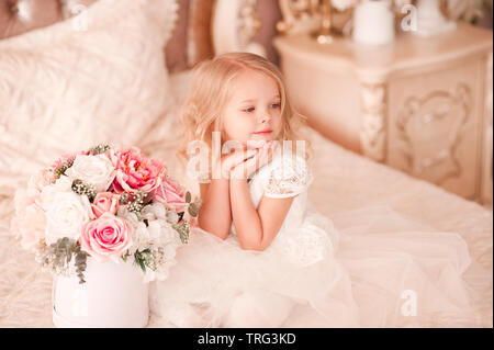 Beautiful baby girl wearing stylish white dress and psong in bed with flowers. Good morning. Happy birthday. Stock Photo