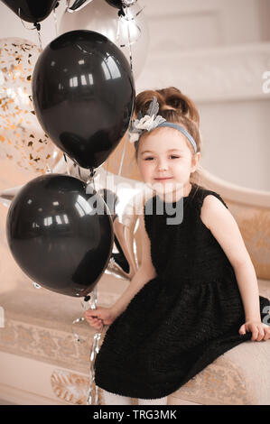 Smiling baby girl 5 6 year old wearing stylish black dress holding balloons sitting on sofa in room. Looking at camera. Celebrating birthday. Childhoo Stock Photo Alamy