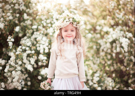 Smiling blonde kid girl holding basket with flowers outdoors. Looking at camera. Wearing floral wreath. Stock Photo