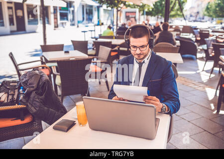 Handsome young man sitting at  street cafe reading documents while working on laptop and wearing headphones. Stock Photo