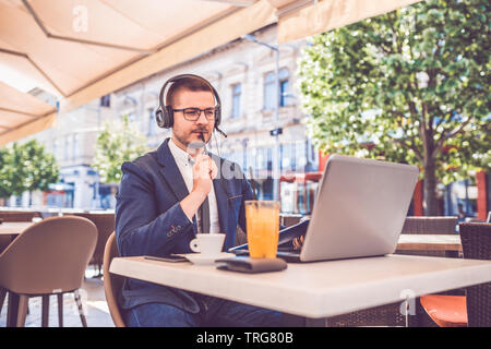 Handsome young man sitting at street cafe reading documents while working on laptop and wearing headphones. Stock Photo