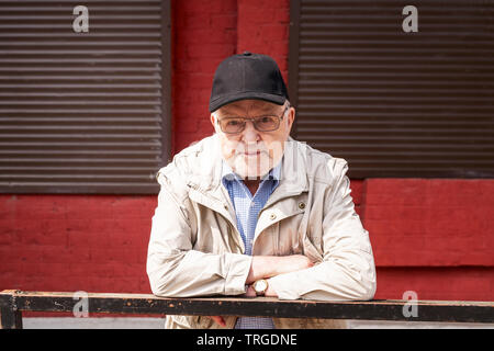 Street portrait of old man in eyeglasses and cap outdoors. Senior posing in front of red wall with windows Stock Photo
