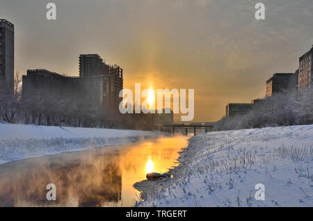 Winter golden sunset and mist rises above the water of little warm river. Frosty weather, trees and grass on the banks are covered with hoarfrost Stock Photo
