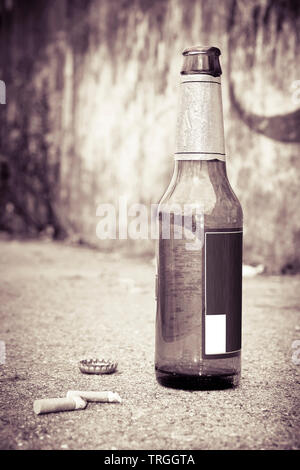 Bottle of beer resting on the ground with three cigarette's butts. Alcoholism and tobacco addiction concept - toned image Stock Photo