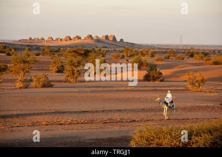 traditionally dressed arab riding a camel in nubian desert. The pyramids of Meroe on the skyline in the early morning sun UNESCO world heritage site Stock Photo