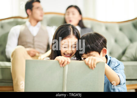 two asian kids brother and sister sitting on carpet reading book together in family living room with parents sitting on couch in the background.
