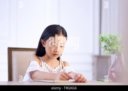 beautiful little asian girl with long black hair sitting at desk in her room writing or drawing on note book Stock Photo