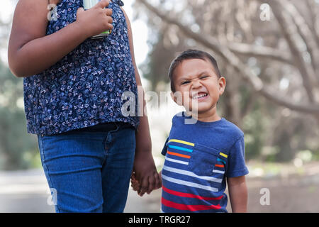 A little boy with colorful shirt with a silly smirk on his face that is holding his older sisters hand. Stock Photo