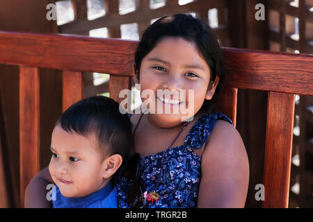 An older sister holding her younger brother tightly while sitting on a wooden bench. Stock Photo