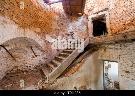 Large spacious forsaken empty basement room of ancient building or palace with cracked plastered brick walls, dirty floor and wooden staircase ladder. Stock Photo