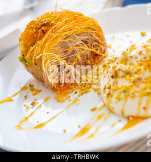 Close up of a succulent Greek dessert. Greek kataifi with honey yogurt and grated pistachios on a white plate and a blurred background. Square view Stock Photo