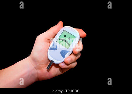 Human hand holding glucometer on black background with sad face on its monitor illustrating bad blood sugar levels. Diabetes concept Stock Photo