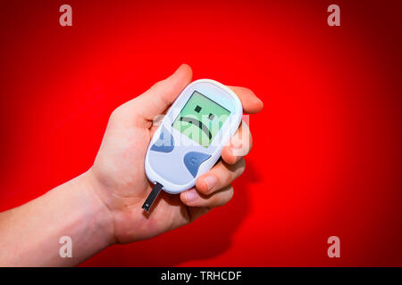 Human hand holding glucometer on red background with sad face on its monitor illustrating bad blood sugar levels. Diabetes concept Stock Photo