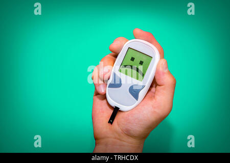 Human hand holding glucometer on turquoise background with sad face on its monitor illustrating bad blood sugar levels. Diabetes concept Stock Photo