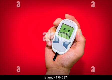 Human hand holding glucometer on red background with sad face on its monitor illustrating bad blood sugar levels. Diabetes concept Stock Photo