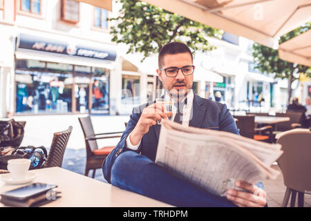 Young businessman with glass of water in hand reading newspaper at outdoor cafe. Stock Photo