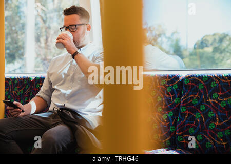 A shot of a mid-adult caucasian businessman sitting on a train commuting to work in Perth, Australia. He is using his smart phone while drinking a hot Stock Photo