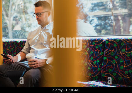 A shot of a mid-adult caucasian businessman sitting on a train commuting to work in Perth, Australia. He is using his smart phone while holding a hot Stock Photo