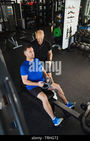 A  high-angle view of a mid-adult caucasian man training in the gym, he is using a rowing machine. A fitness instructor can be seen standing by the ma Stock Photo