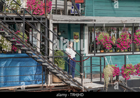 Zemun, Serbia - Young woman standing in the entrance to the raft house restaurant moored at the Danube River shoreline and speaking by smartphone Stock Photo