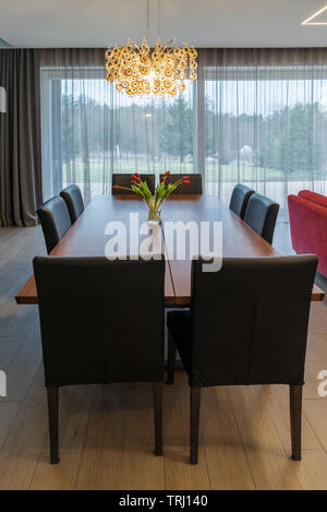 Brown wooden large dining table with chairs in a spacious, bright living room in a private house. The vase with flowers stands on a table Stock Photo