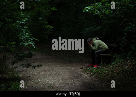 Man sitting on a bench in light spot on forest path, surrounded with darkness, covering his face with his hands, alone, sad and depressed Stock Photo